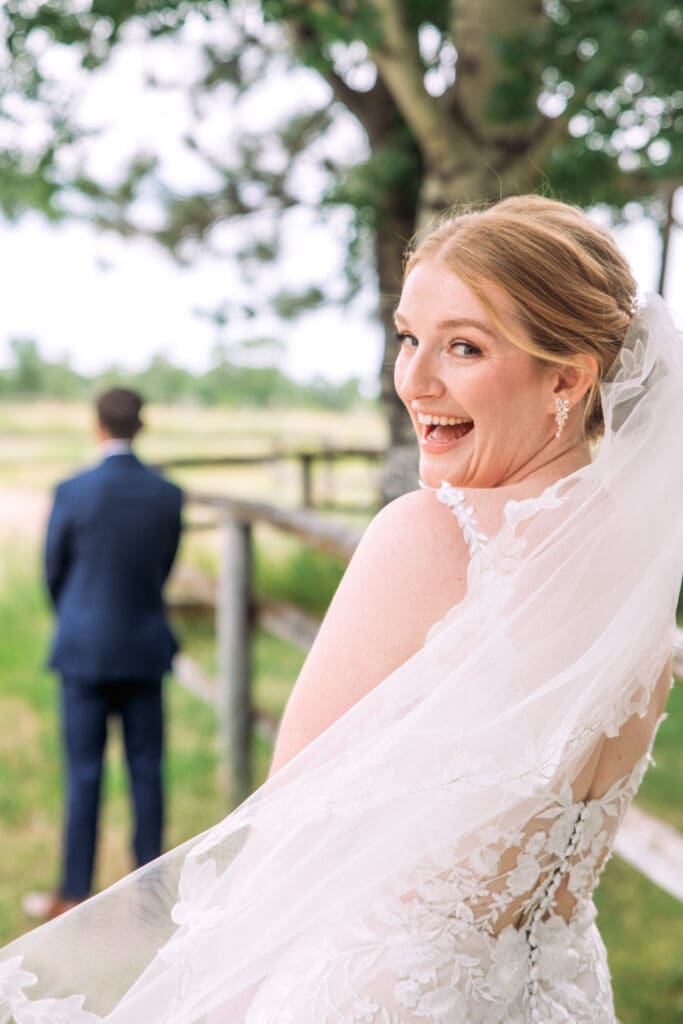Bride turns over her shoulder and smiles widely at camera with groom standing with back to camera in background at the Snake River Meadow wedding venue behind them, captured by Idaho Falls wedding photographer.