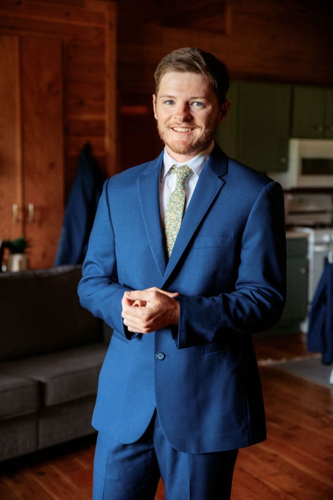 Groom smiles at camera while adjusting blue suit cuffs in wooden groom suite at the Snake River Meadow venue, captured by Idaho Falls wedding photographer.