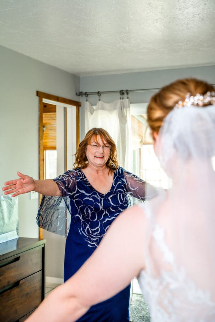 Mom smiles and opens arms to hug bride in bride's suite after doing a first look at the Snake River Meadow venue, captured by Idaho Falls wedding photographer.