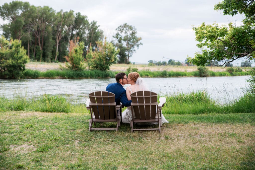 Bride and groom kiss each other while sitting on lawn chairs in front of river at the Snake River Meadow wedding venue, captured by Idaho Falls photographer.