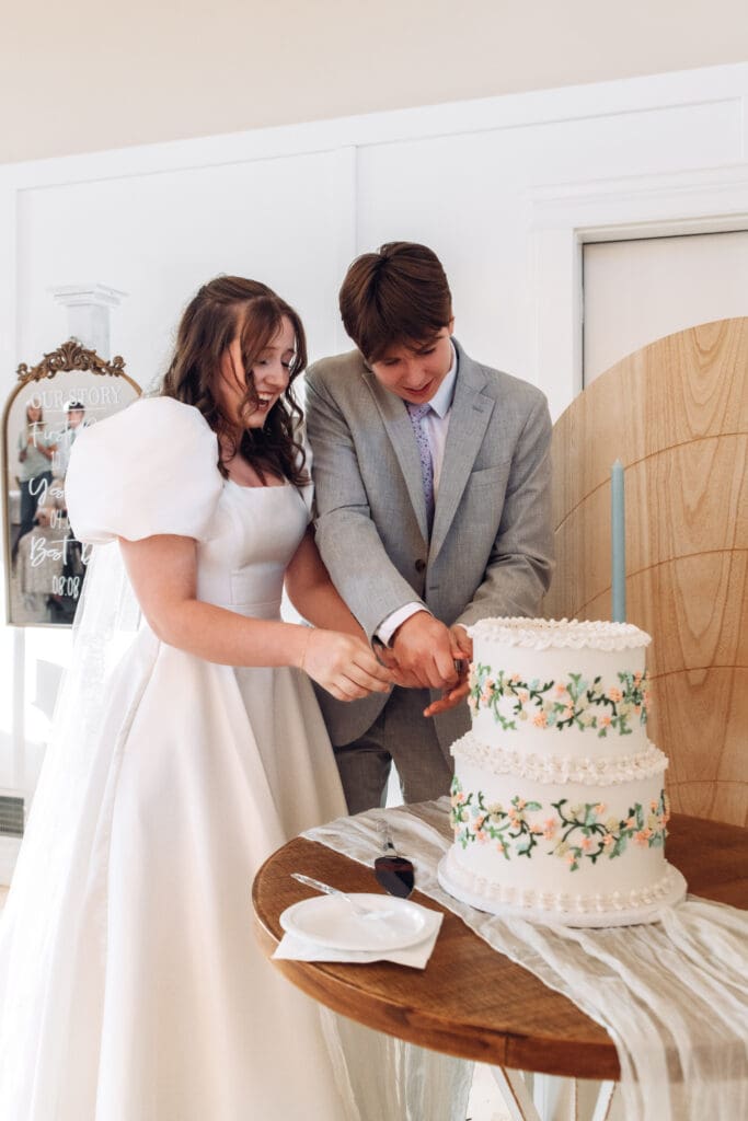 Bride and groom smile as they cut wedding cake together at The Aisle wedding venue, captured by Rexburg wedding photographer. 