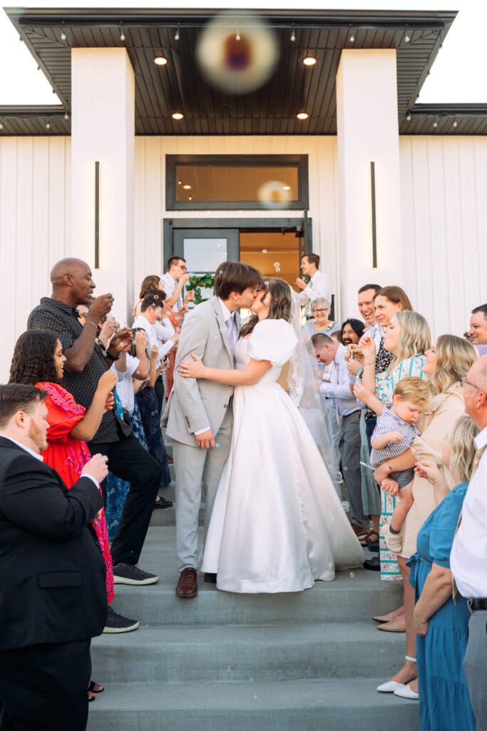Bride and groom kiss on stairs outside The Aisle wedding venue with friends and family clapping, captured by Rexburg wedding photographer. 