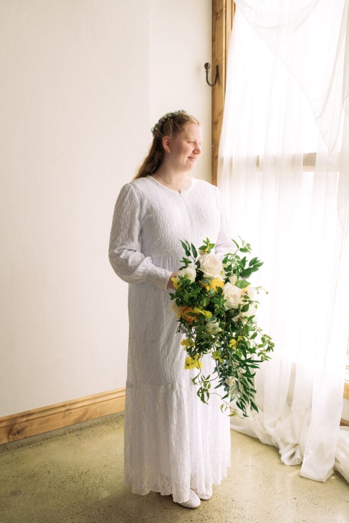 Bride stands in front of window covered with soft curtains while holding long bouquet and smiles at The Atrium, captured by Rexburg wedding photographer.