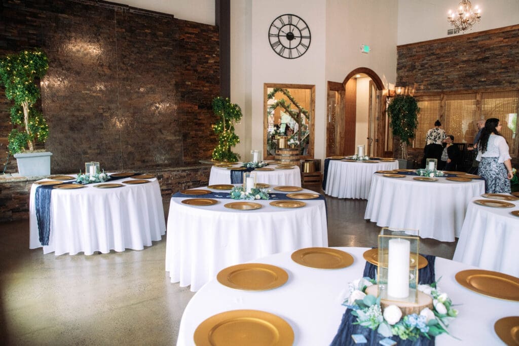 Round tables with white linen, blue cloth runners, candles, and gold plates set up at The Atrium with clock and waterfall in background, captured by Rexburg wedding photographer.
