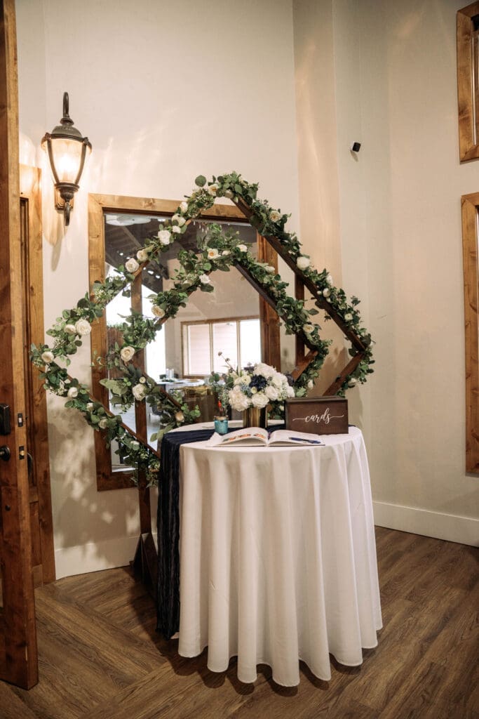 Round guest entrance table with white linen in front of dark wooded diamond arch with greenery at The Atrium, captured by Rexburg wedding photographer.