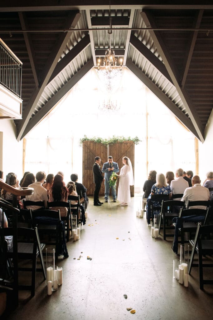 Vaulted ceiling showing off as bride and groom stand in front of officiant and wood backdrop with windows behind them and friends and family watching from black wedding chairs at The Atrium, captured by Rexburg wedding photographer..