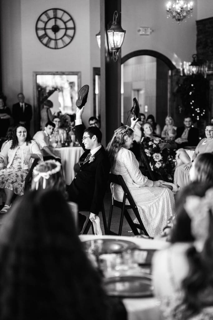 Bride and groom sit in chairs back to back and hold groom's shoes in gamer with family and friends smiling while sitting at round table in back and foregrounds at The Atrium, captured by Rexburg wedding photographer.