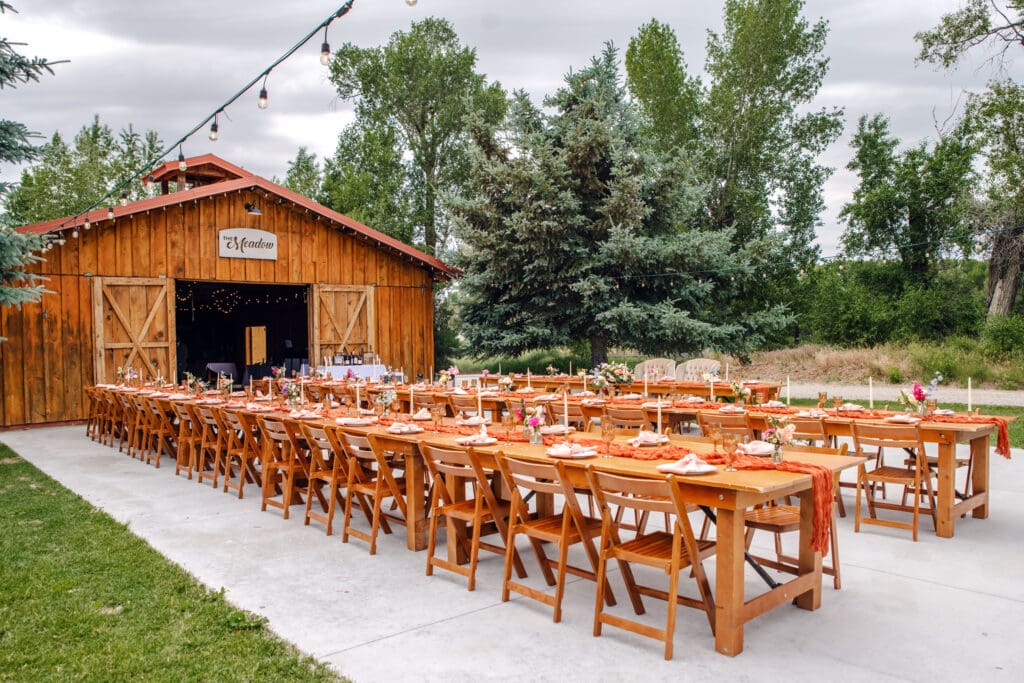 Wooden wedding tables and chairs with orange cloth runner, florals, and candles on concrete pad in front of barn and trees at the Snake River Meadow wedding venue, captured by Idaho Falls wedding photographer.