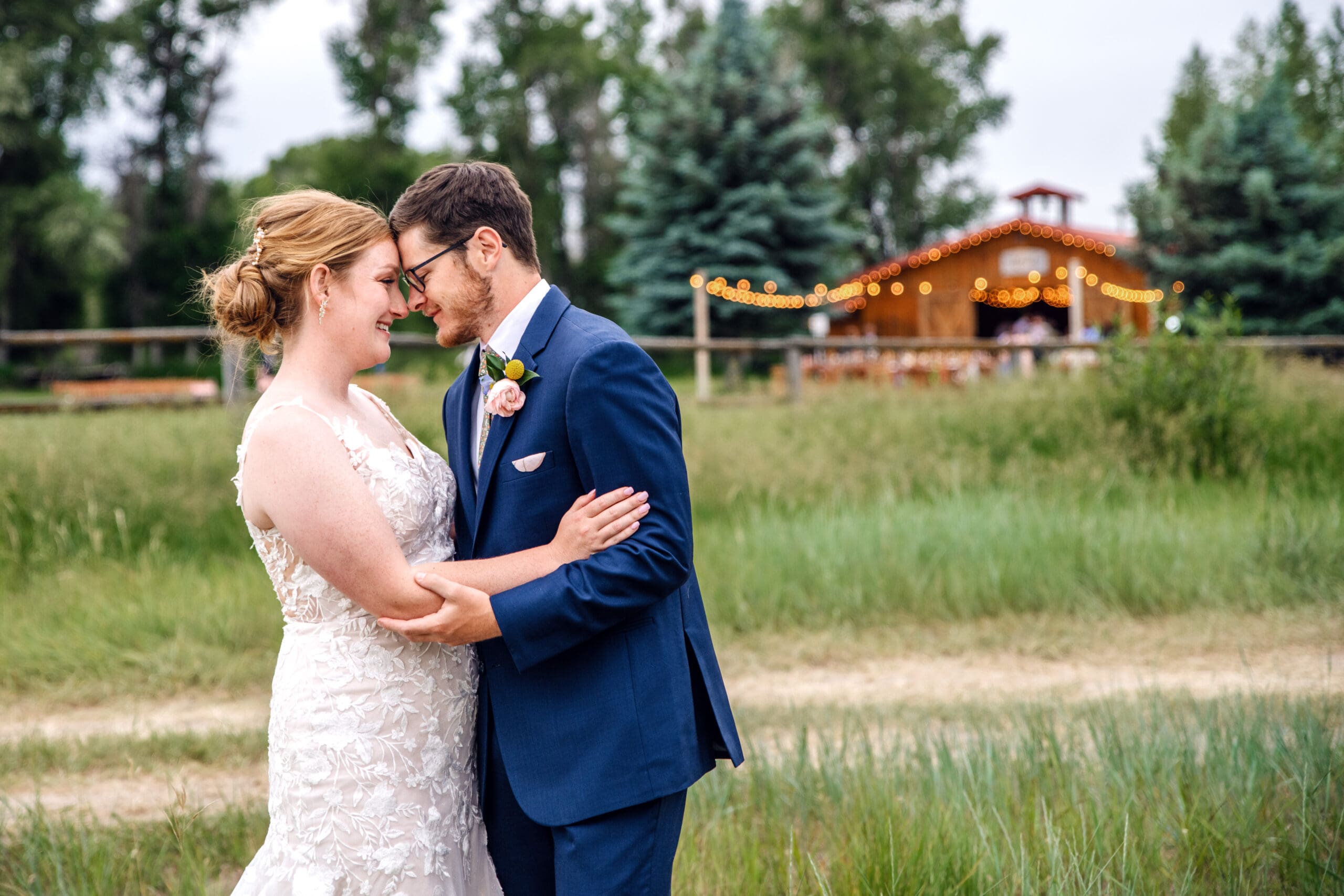 Bride and groom hold each other and press foreheads together while smiling in field of grass with the Snake River Meadow wedding venue behind them, captured by Idaho Falls wedding photographer.