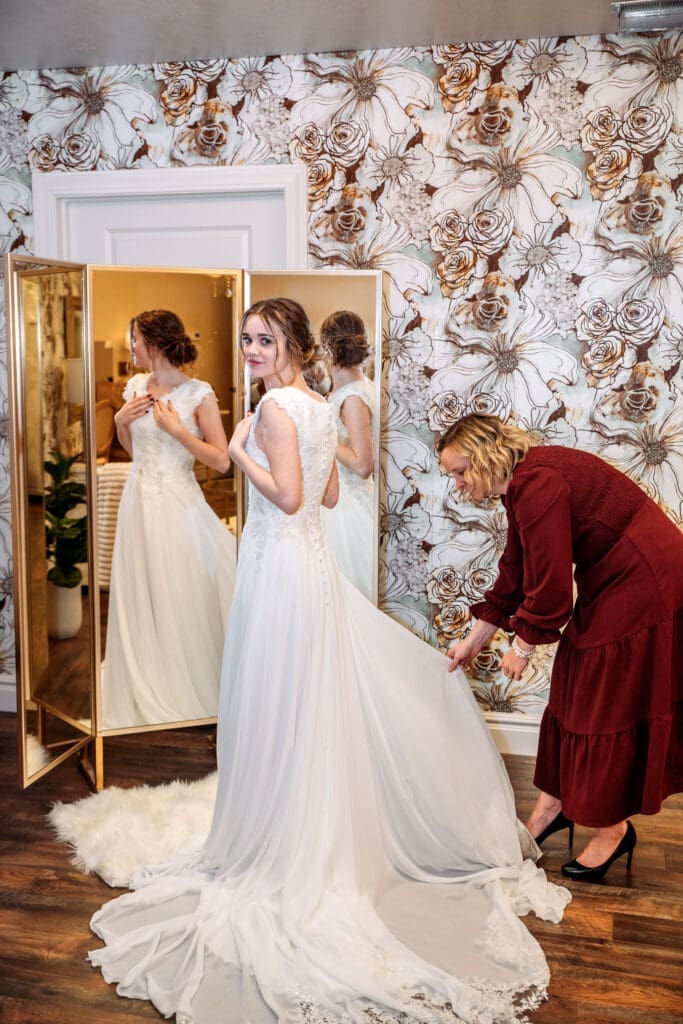 Bride smiles over her shoulder at camera as mom fluffs her dress in front of mirror and decorative wallpaper at The Aisle in Sugar City, captured by Rexburg wedding photographer. 