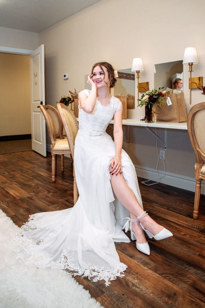 Bride tucks hair behind ear as she smiles and sits in chair in bridal suite with mirrors behind her at The Aisle wedding venue, captured by Rexburg wedding photographer. 