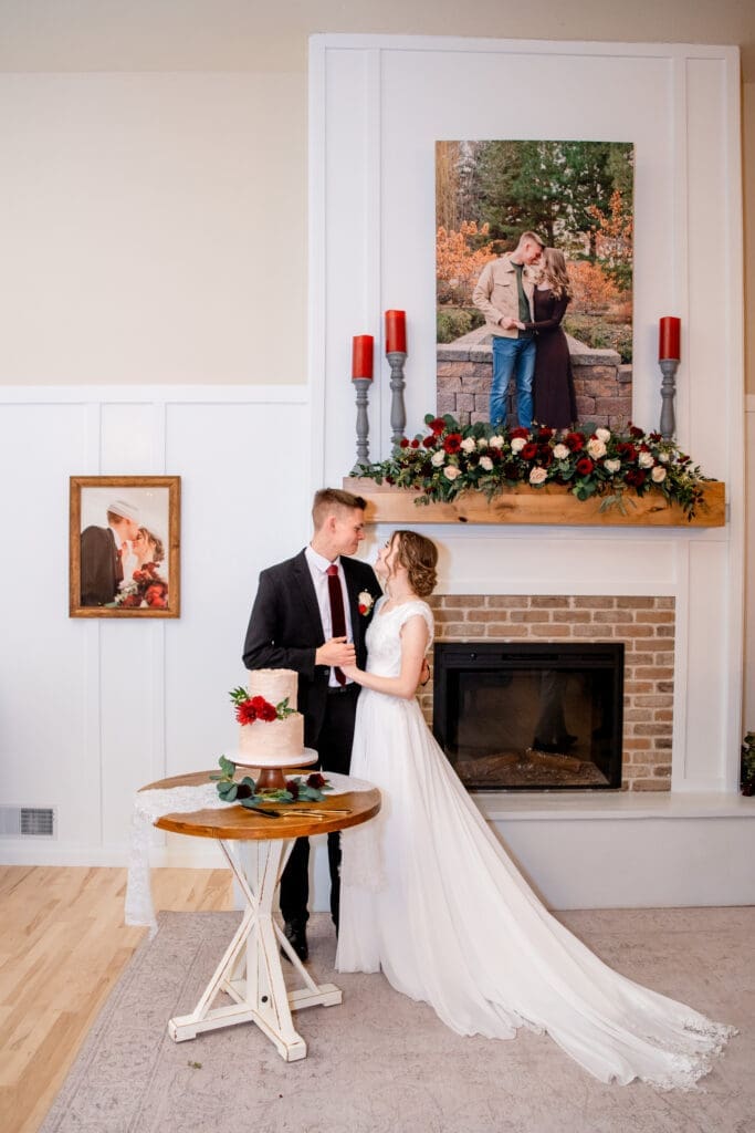Bride and groom hold hands and smile at each other and hold hands in front of wedding cake with fireplace and photos behind them at The Aisle wedding venue, captured by Rexburg wedding photographer. 