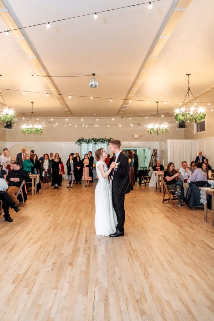 Bride and groom smile up at each other during their first dance with family and friends watching at The Aisle wedding venue, captured by Rexburg wedding photographer. 