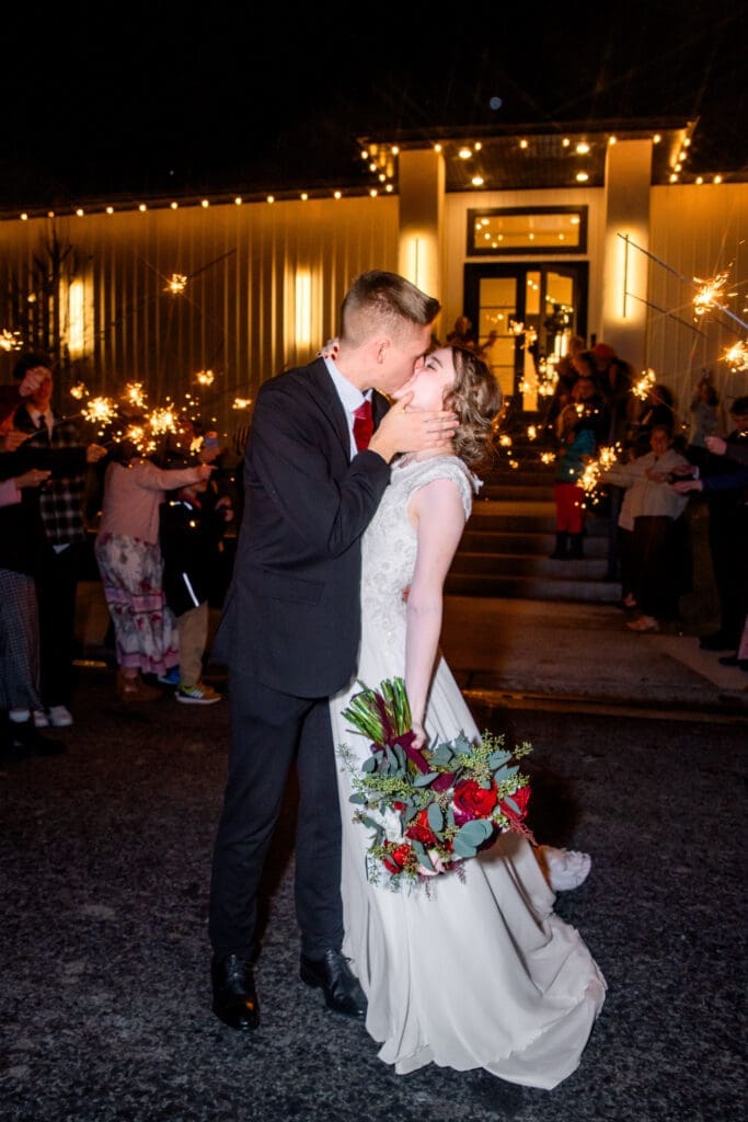 Bride and groom kiss with bride holding flowers and groom placing hand on her jaw outside The Aisle wedding venue with friends and family holding sparklers, captured by Rexburg wedding photographer. 