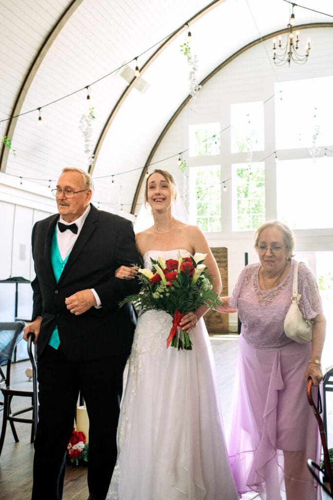 Bride smiles and walks down aisle with dad in blue and black suit and mom in purple dress and barn white barn interior and windows behind them at The Barn on 1st venue, captured by Idaho Falls wedding photographer.
