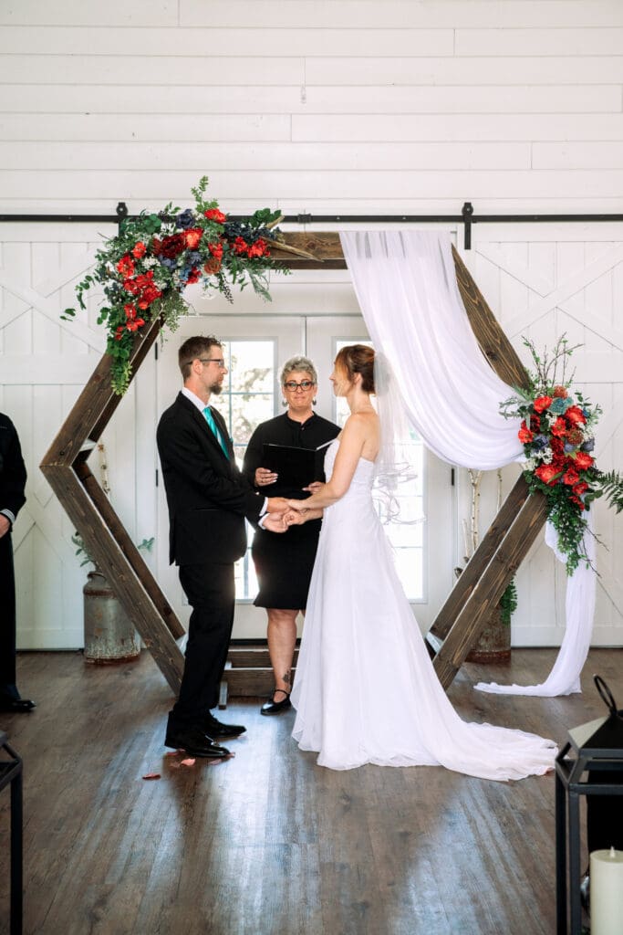 Groom places hands on bride's hips and kisses her cheek from behind as bride holds red and white bouquet in front of stairs and white barn, at The Barn on 1st venue, captured by Idaho Falls wedding photographer.