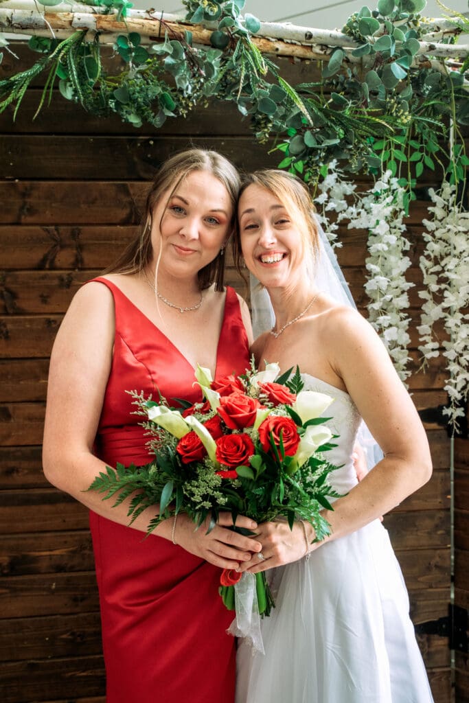 Bride and bridesmaid in red dress smile at camera and hold wedding bouquet with wood shiplap backdrop at The Barn on 1st venue, captured by Idaho Falls wedding photographer. 