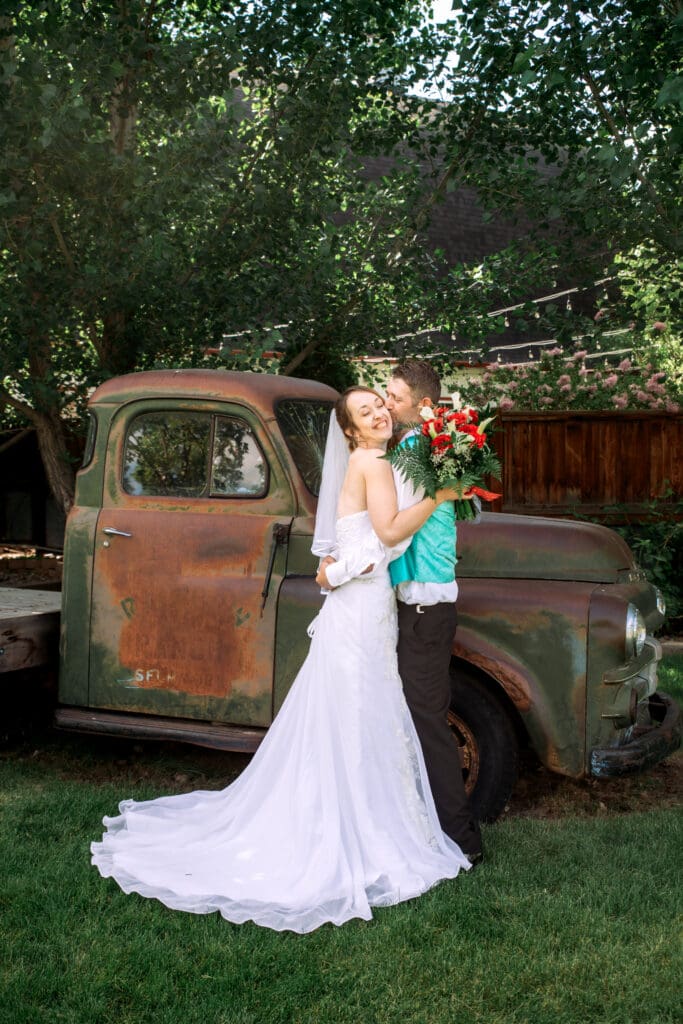 Bride and groom hug as groom kisses bride's cheek and bride smiles and hold bouquet by his head on grass in front of vintage trick at The Barn on 1st, captured by Idaho Falls wedding photographer.