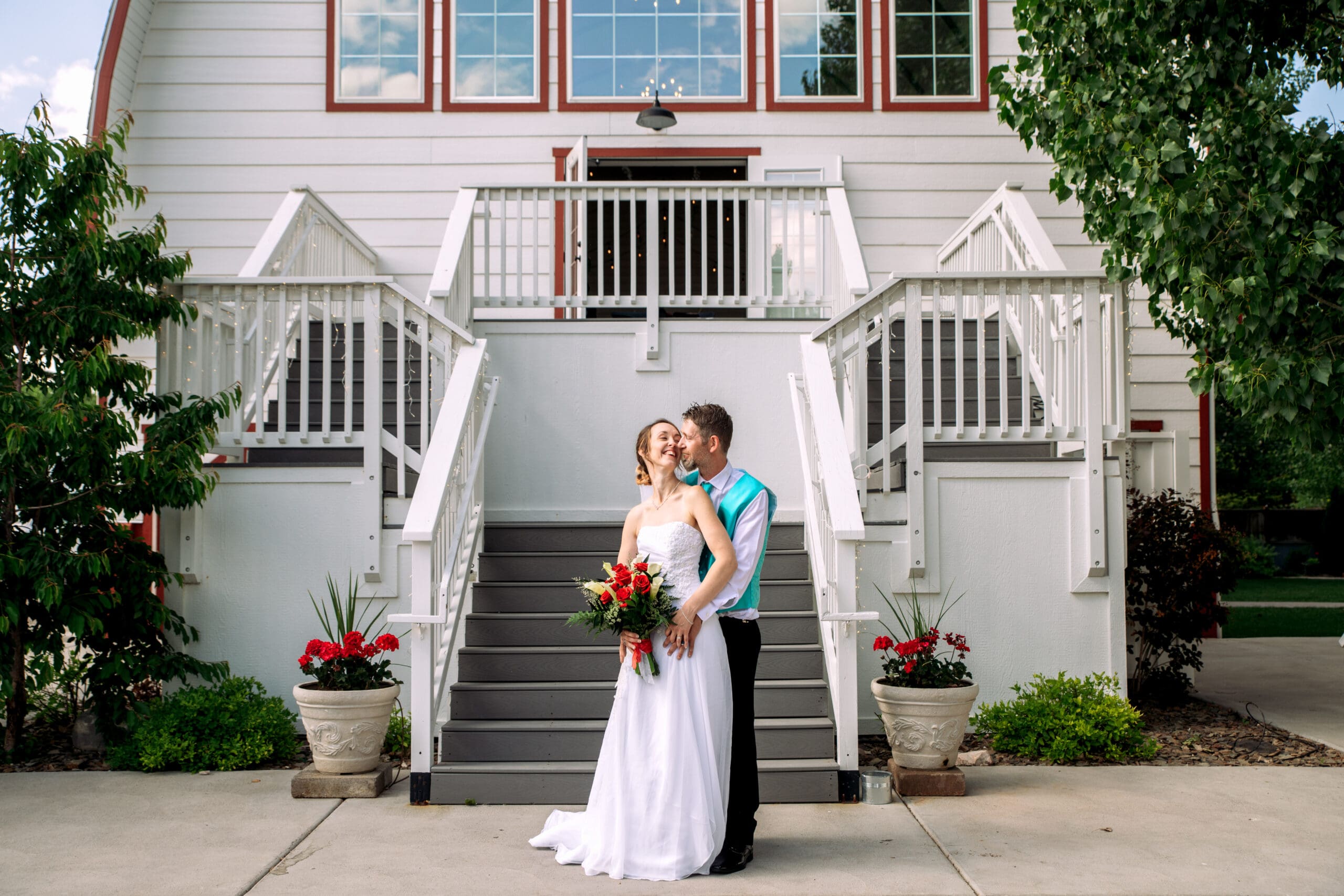 Groom places hands on bride's hips and kisses her cheek from behind as bride holds red and white bouquet in front of stairs and white barn, at The Barn on 1st venue, captured by Idaho Falls wedding photographer.