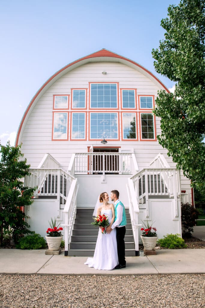 Groom places hands on bride's hips and smiles at her from behind as bride holds red and white bouquet in front of stairs and white barn and smiles back, at The Barn on 1st venue, captured by Idaho Falls wedding photographer.