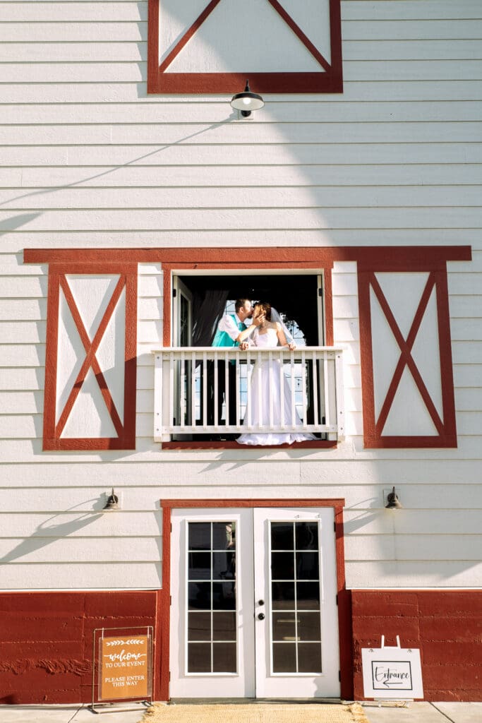 Groom and bride stand on balcony at red and white barn as bride holds rail and groom kiss her while holding her cheek at the Barn on 1st venue, captured by Idaho Falls wedding photographer.
