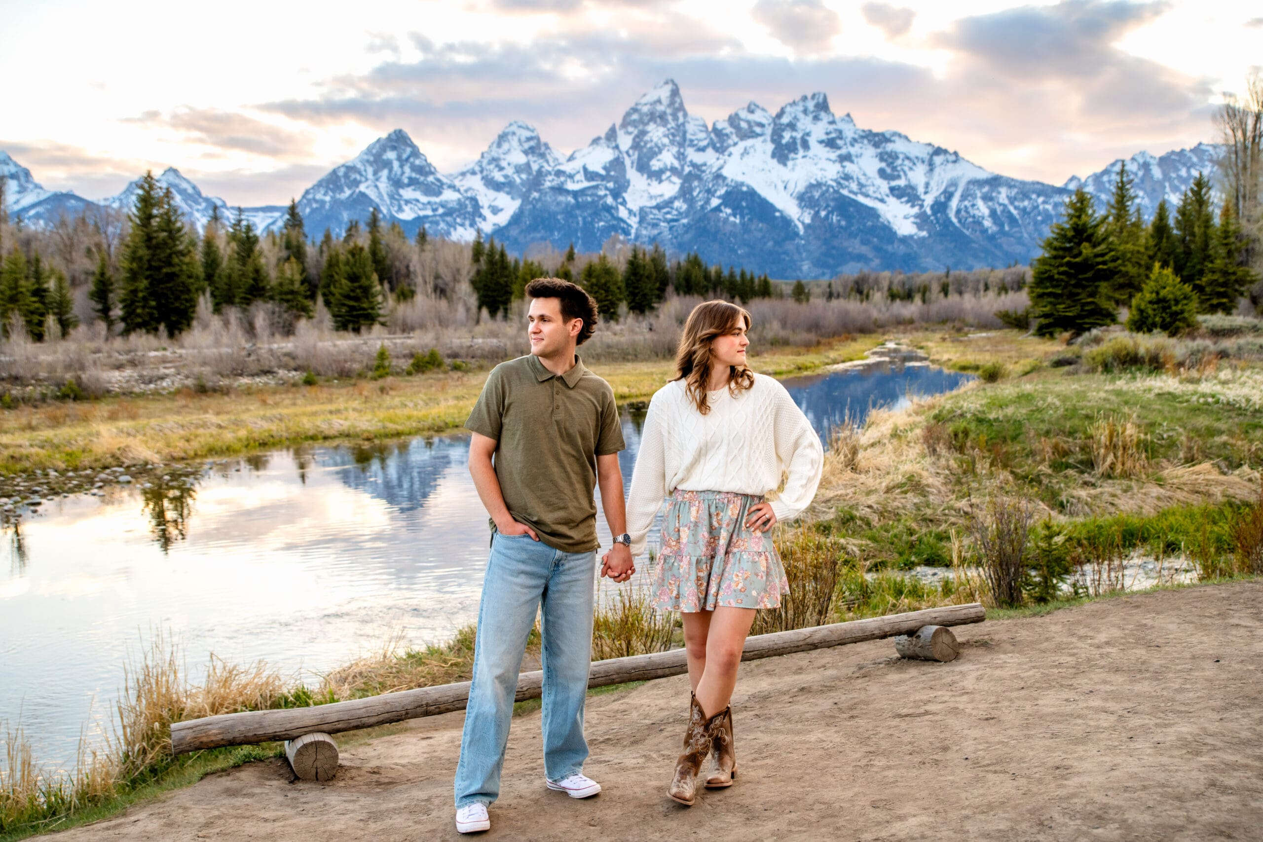 Man and woman stand next to each other while holding hands and looking away over their shoulders in front of mountains and river, captured by Grand Teton engagement photographer.